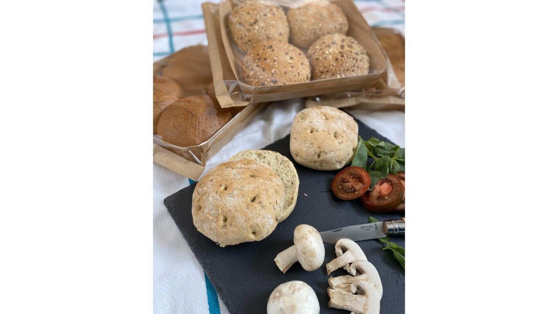 Eine Schieferplatte mit Kräuterbrötchen, geschnittenen Pilzen, Tomaten und Grünzeug. Im Hintergrund stehen auf einem karierten Tuch zwei Tabletts mit verschiedenen Brötchen, die teilweise mit Körnern belegt sind.