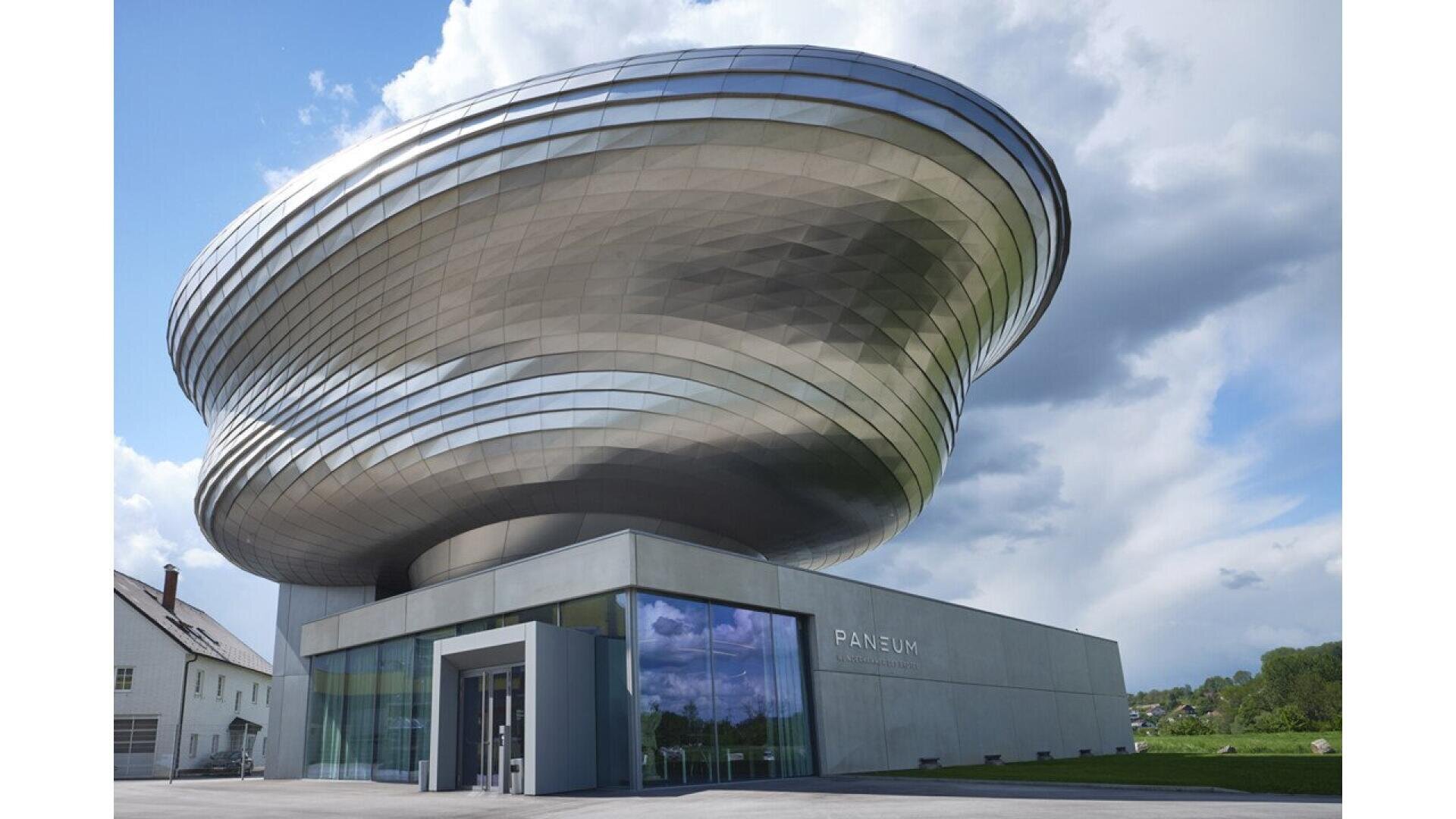 A modern building with a large, metallic, spiraling upper structure and glass-walled ground floor labeled PANEUM, set against a partly cloudy sky and green landscape.
