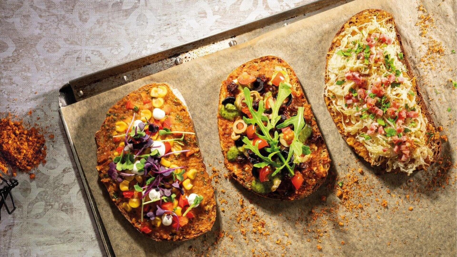 Three flatbread pizzas with assorted colorful toppings, including vegetables, greens, and herbs, are arranged on a parchment-lined baking tray; crumbs are scattered on the side.