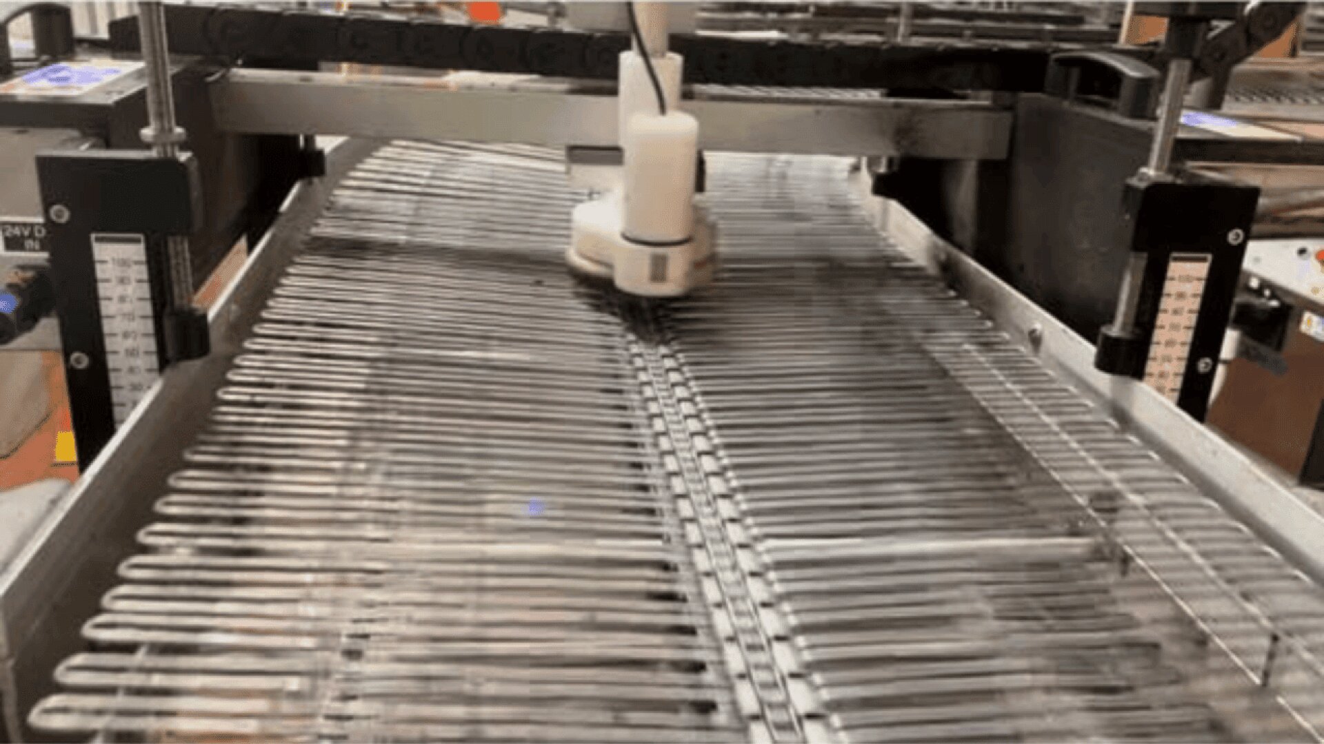 A robotic arm arranges multiple glass laboratory test tubes in rows on a metal conveyor inside an industrial facility, likely part of an automated sorting or cleaning process.