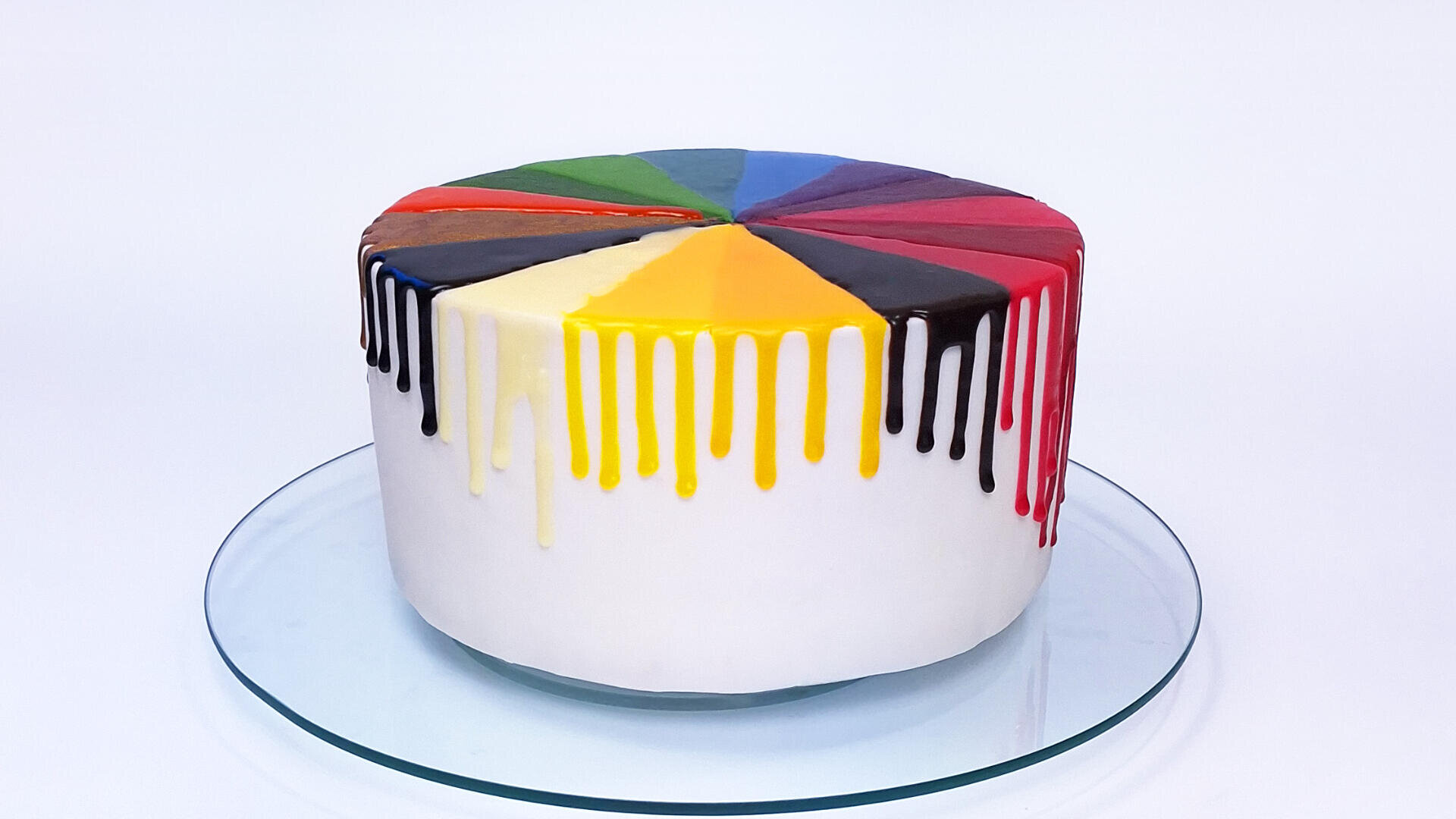 A round cake with white frosting, topped with colorful drips in wedge-shaped sections, each a different color like yellow, red, black, blue, green, and brown, sitting on a glass plate against a white background.