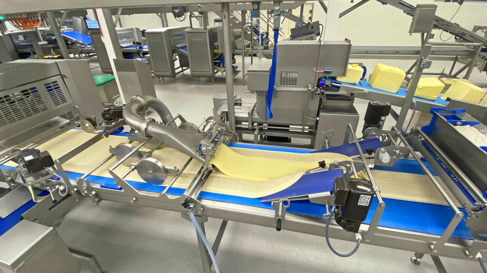 A food processing factory with machines and conveyor belts handling sheets of dough, surrounded by equipment and blocks of yellow butter or margarine in a clean, industrial environment.