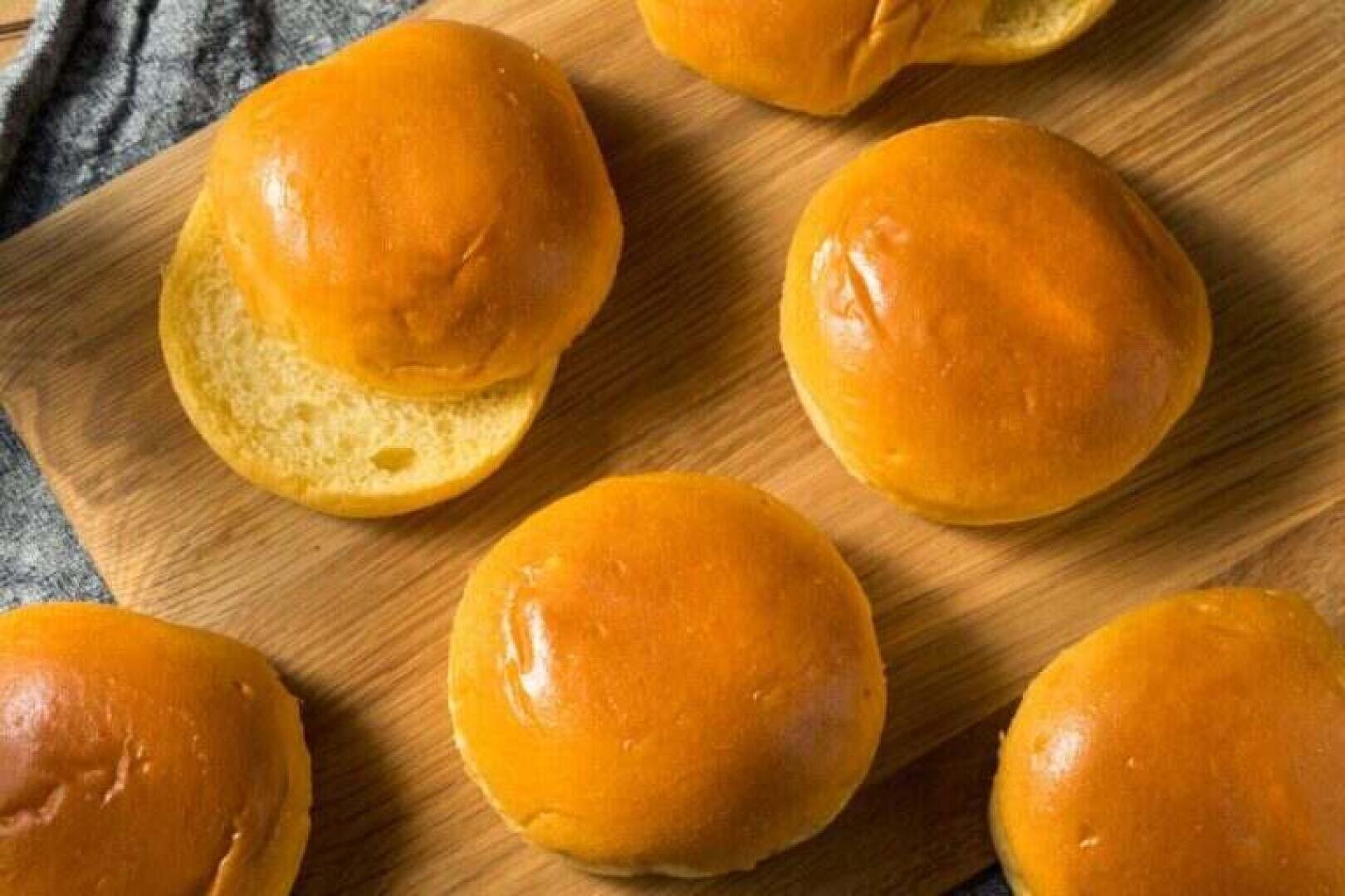 Six golden brown brioche buns are arranged on a wooden base, with one bun partially opened to reveal its soft, fluffy interior. The surface and texture of the buns look shiny and fresh.