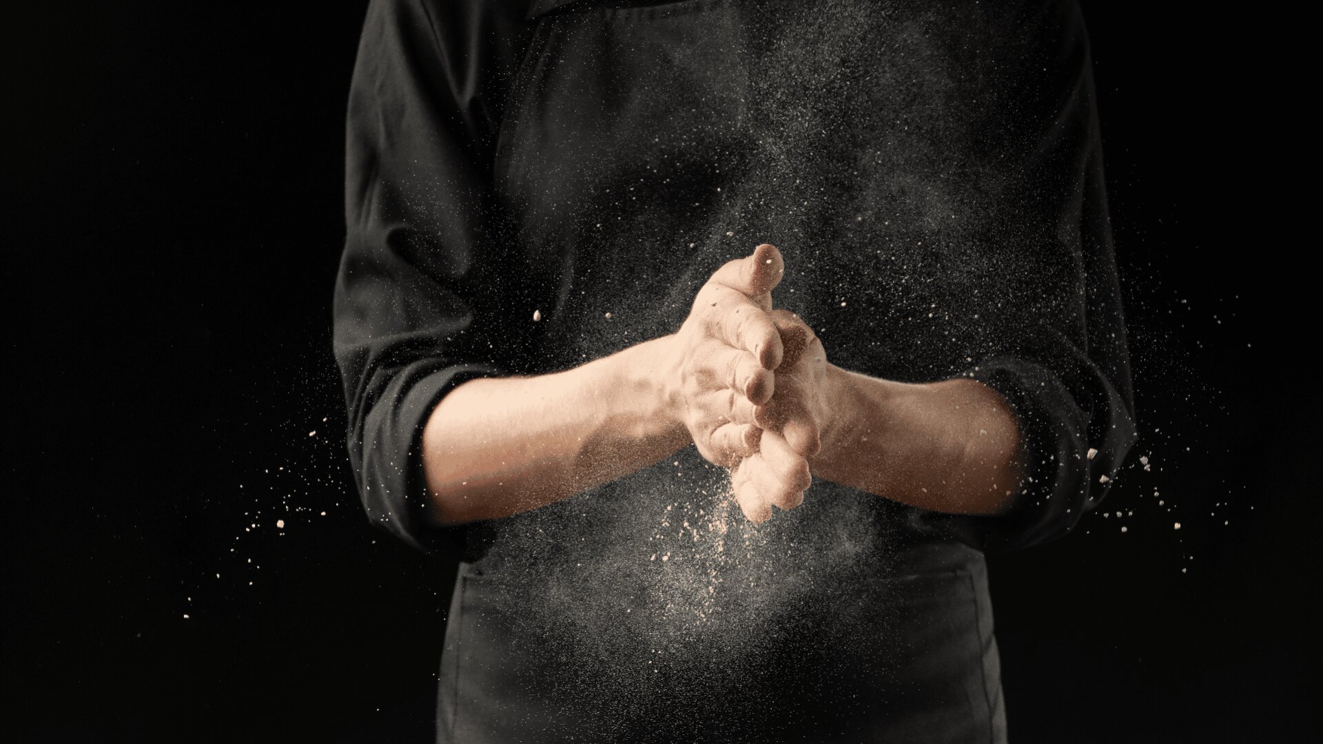 Eine Person in einer schwarzen Schürze klatscht in die Hände und erzeugt eine Mehlwolke, die vor einem dunklen Hintergrund in der Luft schwebt.