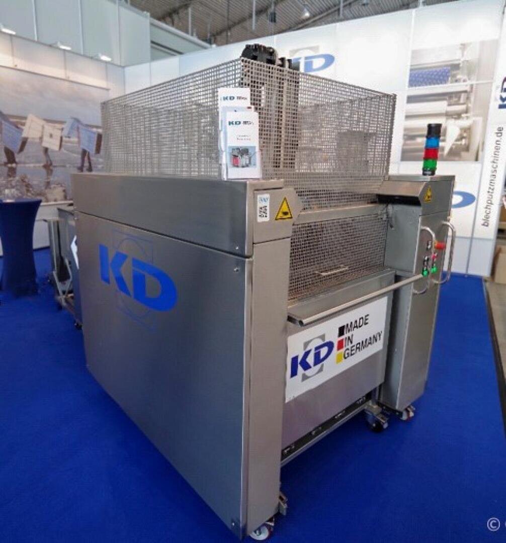 Eine große Industriemaschine mit einem Metallgittergehäuse steht auf einem blauen Teppich; sie zeigt das KD-Logo, "MADE IN GERMANY", Bedienknöpfe und Warnschilder. Im Hintergrund sind Messestände zu sehen.