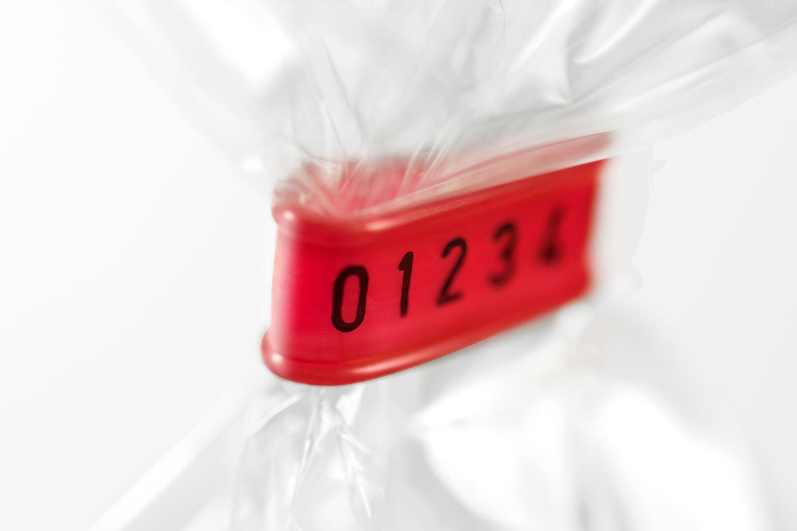 Close-up of a red plastic tag with the numbers 01234 printed in black, fastened around clear plastic, possibly sealing a bag. The background is plain and out of focus.