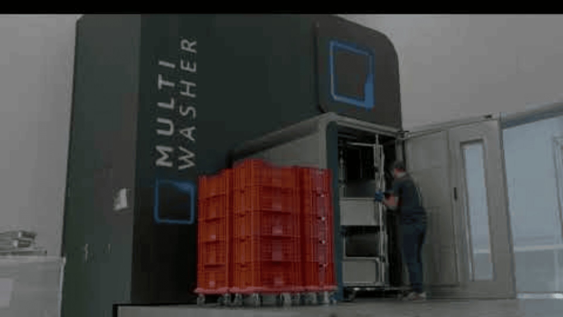 Eine Person lädt rote Plastikkisten in eine große Industriemaschine mit der Aufschrift „Multi Washer“ in einer sauberen, modernen Anlage.