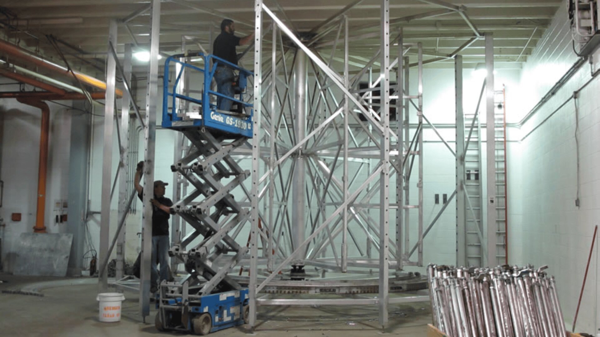 Two workers assemble a large metal structure indoors; one stands on a scissor lift, the other on the floor. Metal beams and construction equipment are scattered around the well-lit industrial space.