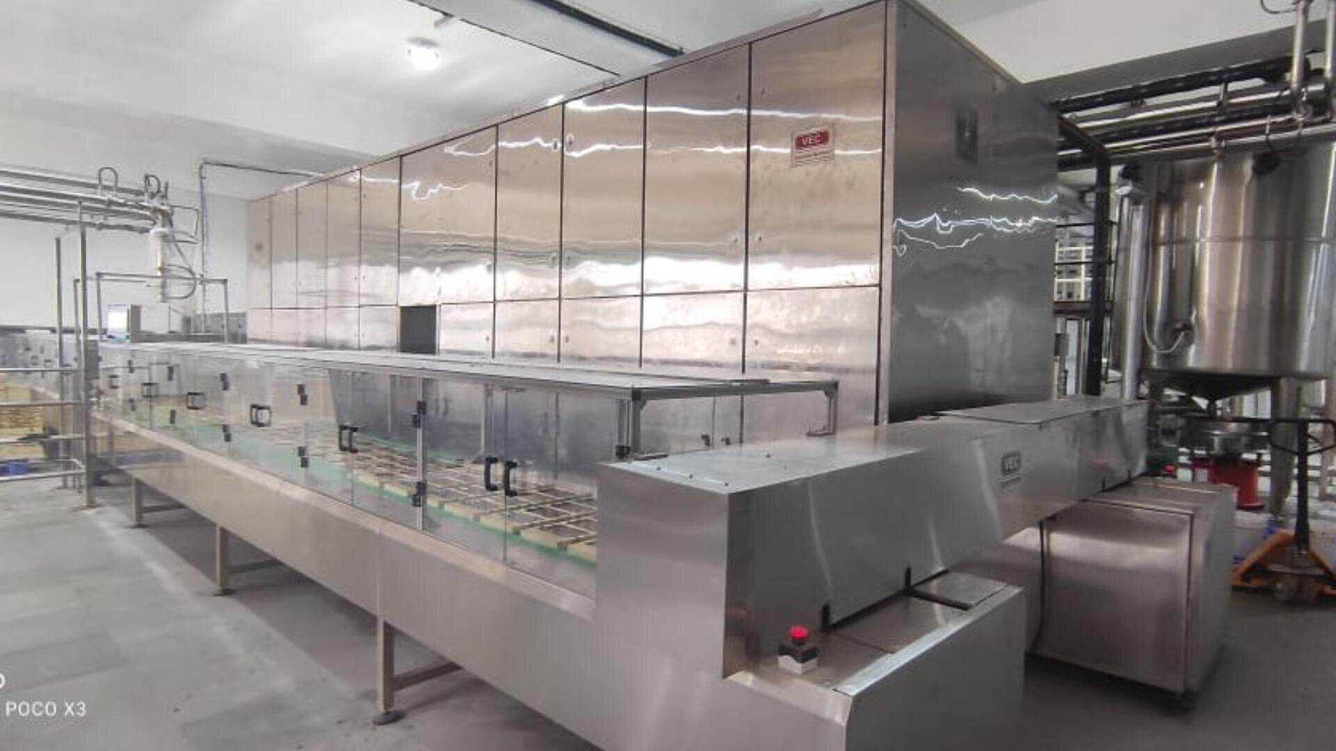 A large, stainless steel industrial bottling or packaging machine stands in a clean, well-lit factory setting, surrounded by pipes and other equipment.