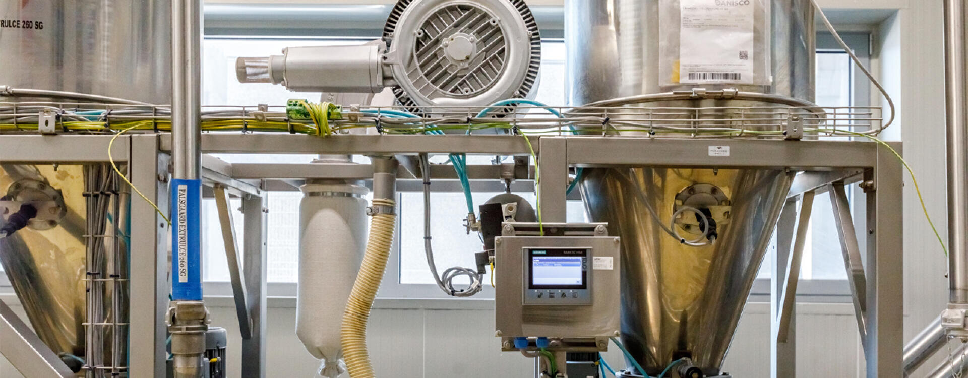 Industrial equipment featuring large stainless steel tanks, pipes, wires, and a control panel in a clean, well-lit facility; machinery is used for processing or manufacturing purposes.