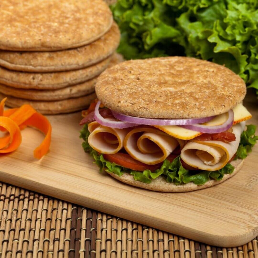 A sandwich made with round whole grain flatbread, rolled turkey slices, cheese, lettuce, tomato, bacon, and raw onion sits on a wooden board, with leafy lettuce and flatbreads in the background.
