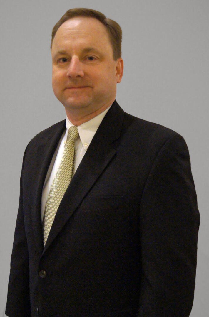 Ein Mann in einem dunklen Anzug, hellem Hemd und gemusterter Krawatte steht vor einem schlichten hellgrauen Hintergrund und blickt mit neutralem Blick in die Kamera.