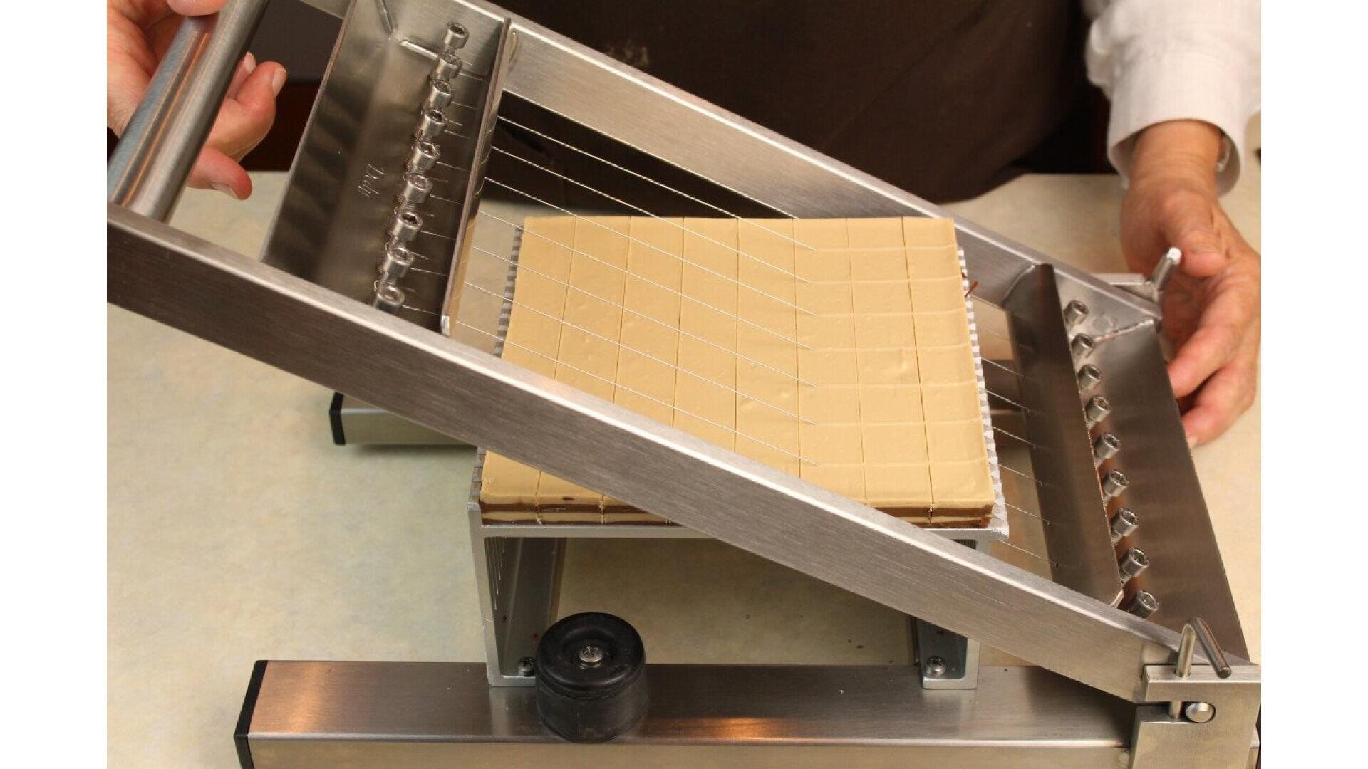 A person uses a metal wire slicing machine to cut a rectangular block of beige-colored material, possibly chocolate or nougat, into even pieces on a countertop.