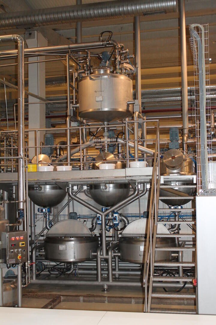 Große Industrietanks und Rohrleitungen aus Edelstahl in einer Lebensmittel- oder Chemieanlage, mit Schalttafel, Treppe und mehreren miteinander verbundenen Behältern und Maschinen.