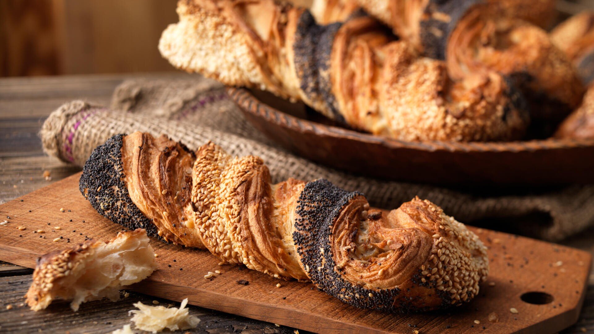 Eine Nahaufnahme eines gedrehten, goldbraunen Gebäckstücks, bestreut mit Sesam und Mohn, liegt auf einem Holzschneidebrett, daneben liegen Krümel. Im Hintergrund stapeln sich weitere Gebäckstücke in einer rustikalen Schüssel.
