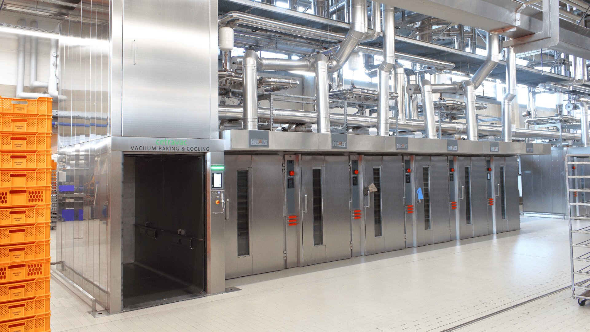 A row of large stainless steel industrial ovens with control panels stands in a clean factory setting, surrounded by metal pipes and orange plastic crates on the left.