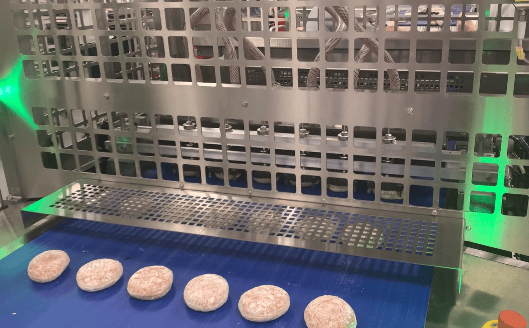Six round, breaded food items are lined up on a blue conveyor belt under a large, metal industrial machine with square cutouts, likely used for food processing in a factory setting.