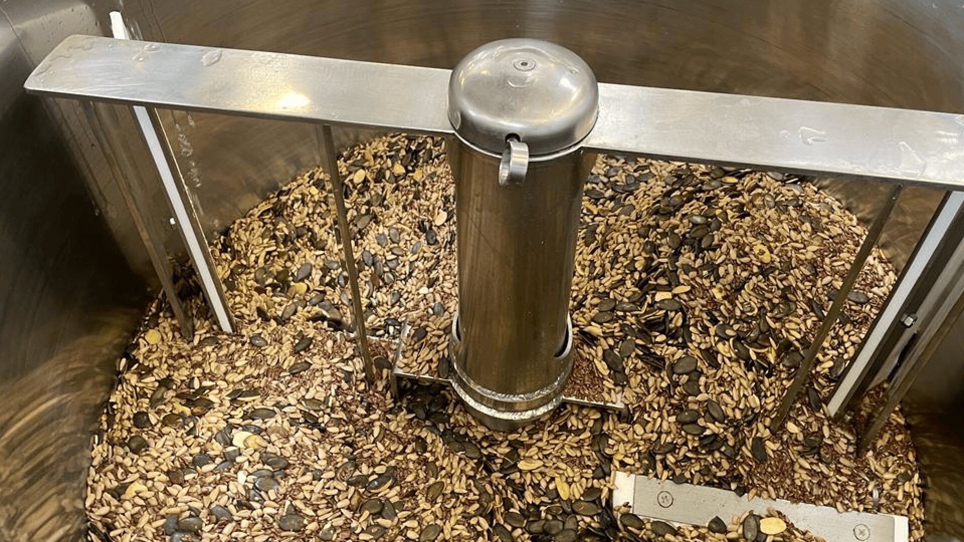 Eine Metallmischmaschine ist mit verschiedenen Samen, darunter Kürbis- und Sonnenblumenkerne, gefüllt, die in einem großen Edelstahlbehälter miteinander verrührt werden.