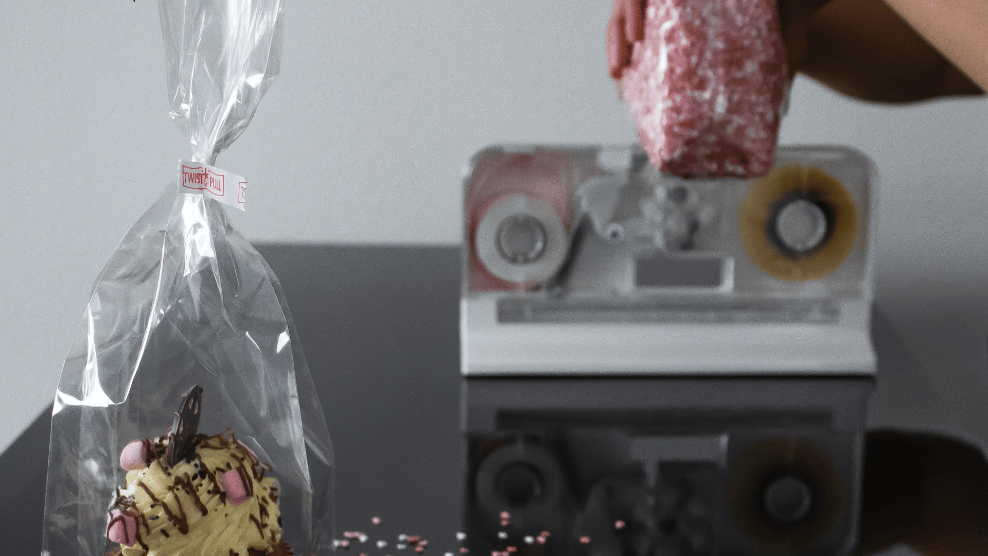 A hand wraps a pink sprinkled treat in plastic film near a dispenser, while a decorated cupcake in a clear bag sits on a reflective black surface.