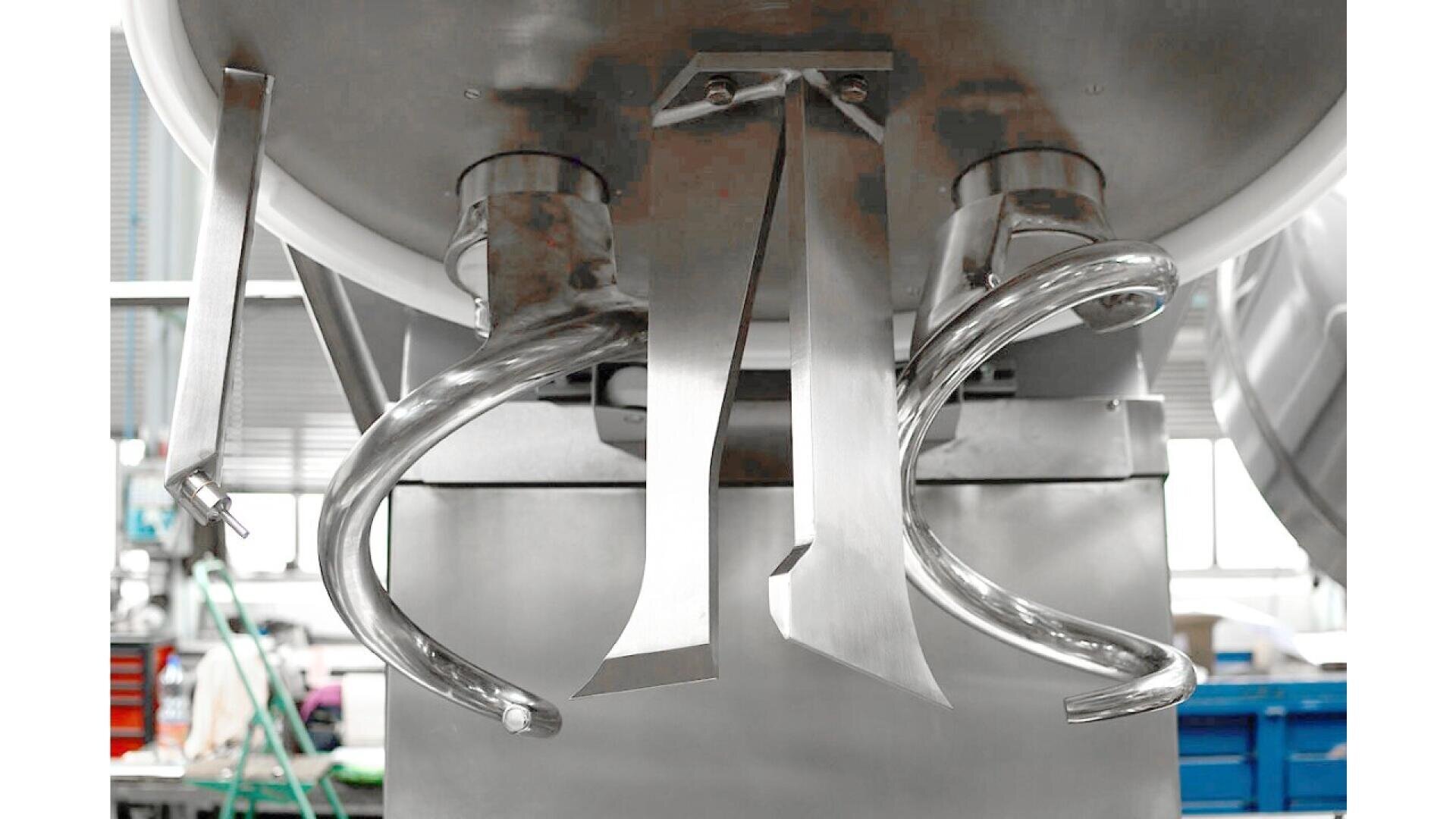 Nahaufnahme von industriellen Mischmaschinenaufsätzen, einschließlich spiral- und klingenförmiger Metallwerkzeuge, die an der Unterseite einer großen Edelstahlmischschüssel in einer Fabrik oder Produktionsumgebung angebracht sind.
