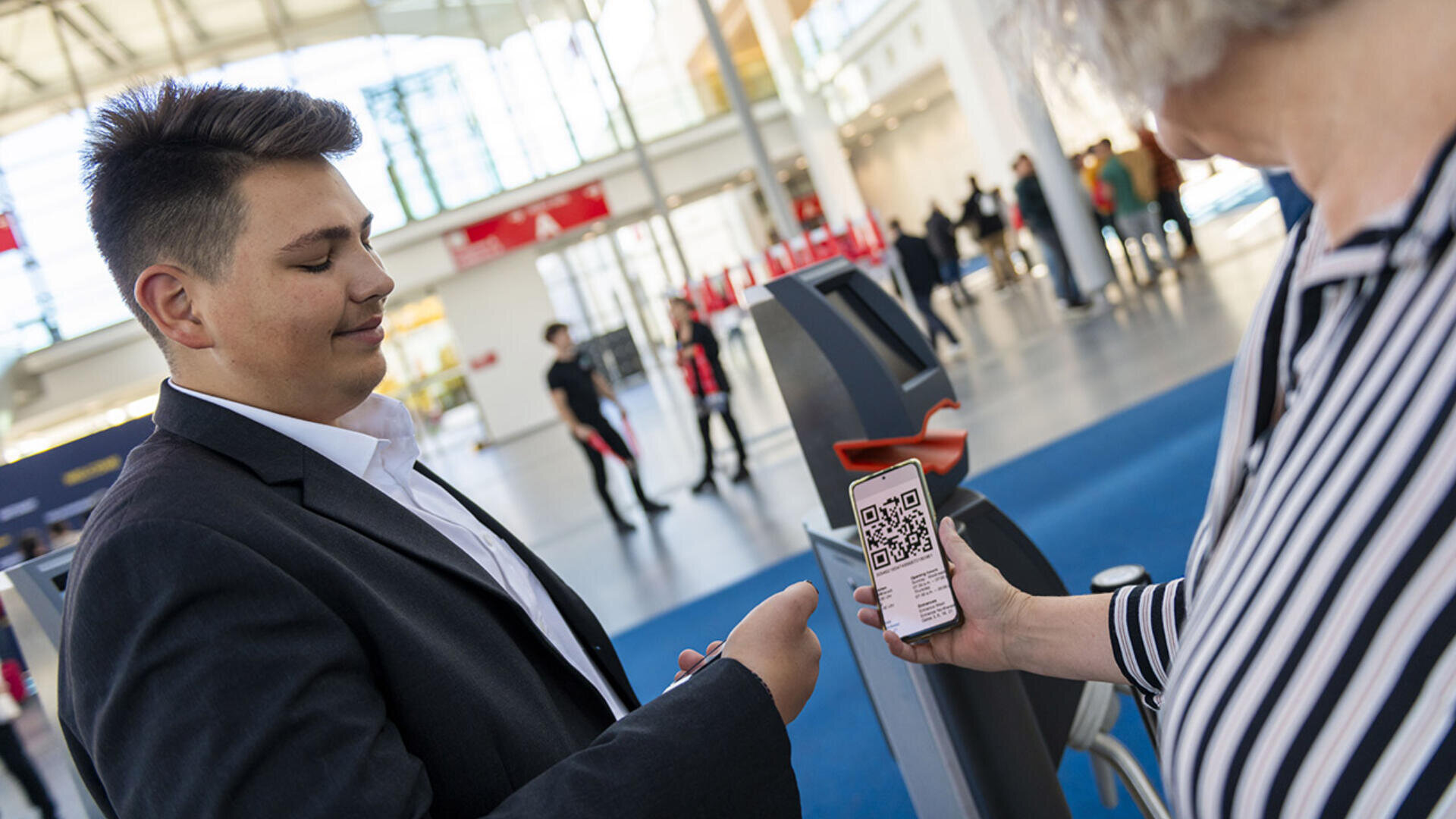 A man in a suit scans a QR code from a phone held by a person in a striped shirt, inside a bright, modern building with people and kiosks in the background.