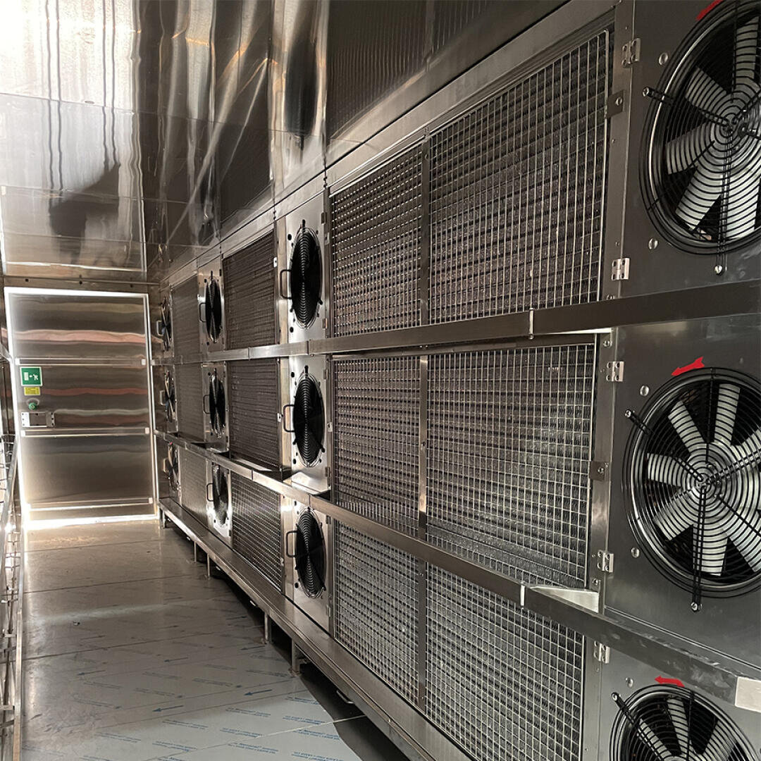 A row of large industrial fans installed behind metal grilles lines the right wall of a metallic, well-lit corridor, leading to a closed door at the end.