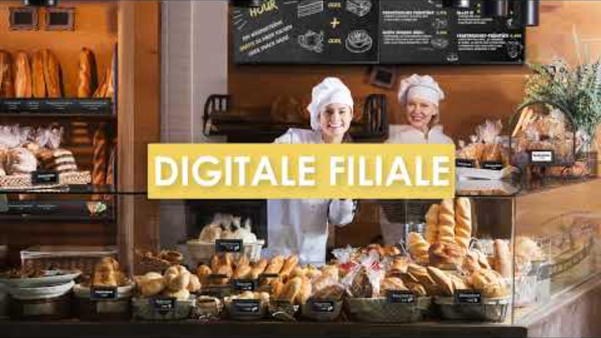 Two bakers in white uniforms and hats stand smiling behind a bakery counter filled with bread and pastries. The text DIGITALE FILIALE appears in yellow across the image.