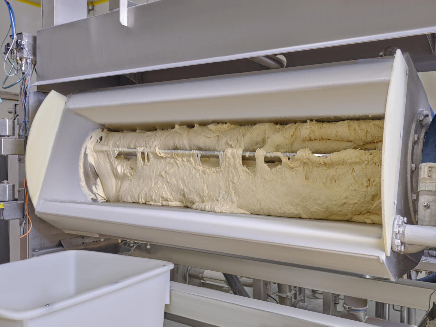 A large industrial dough mixer with freshly mixed dough inside, showing rotating blades and a metal container in a food processing facility.