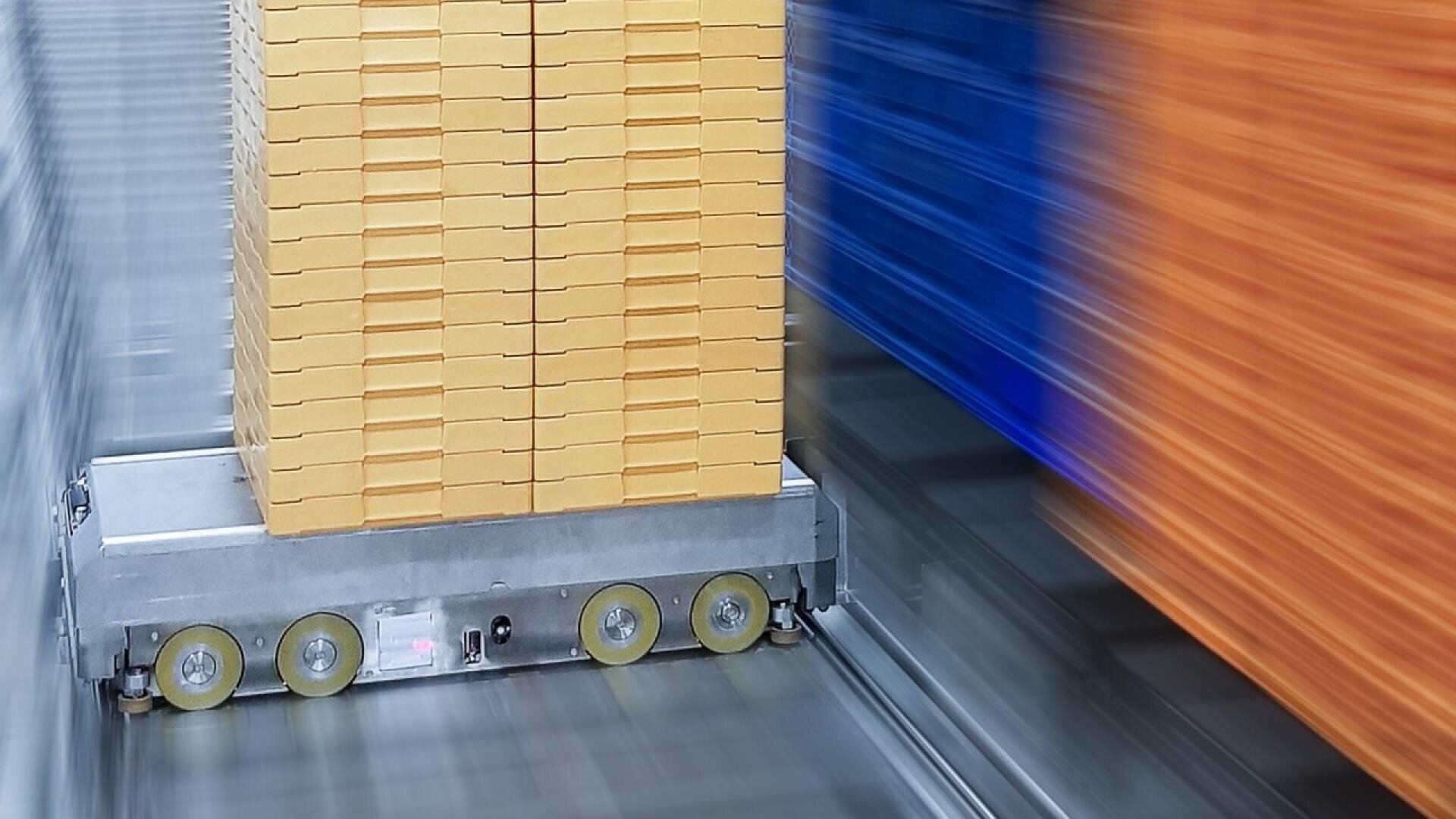 A robotic cart transports a tall stack of yellow boxes through a warehouse, with motion blur indicating speed; blue and orange shelves are visible on the right side.