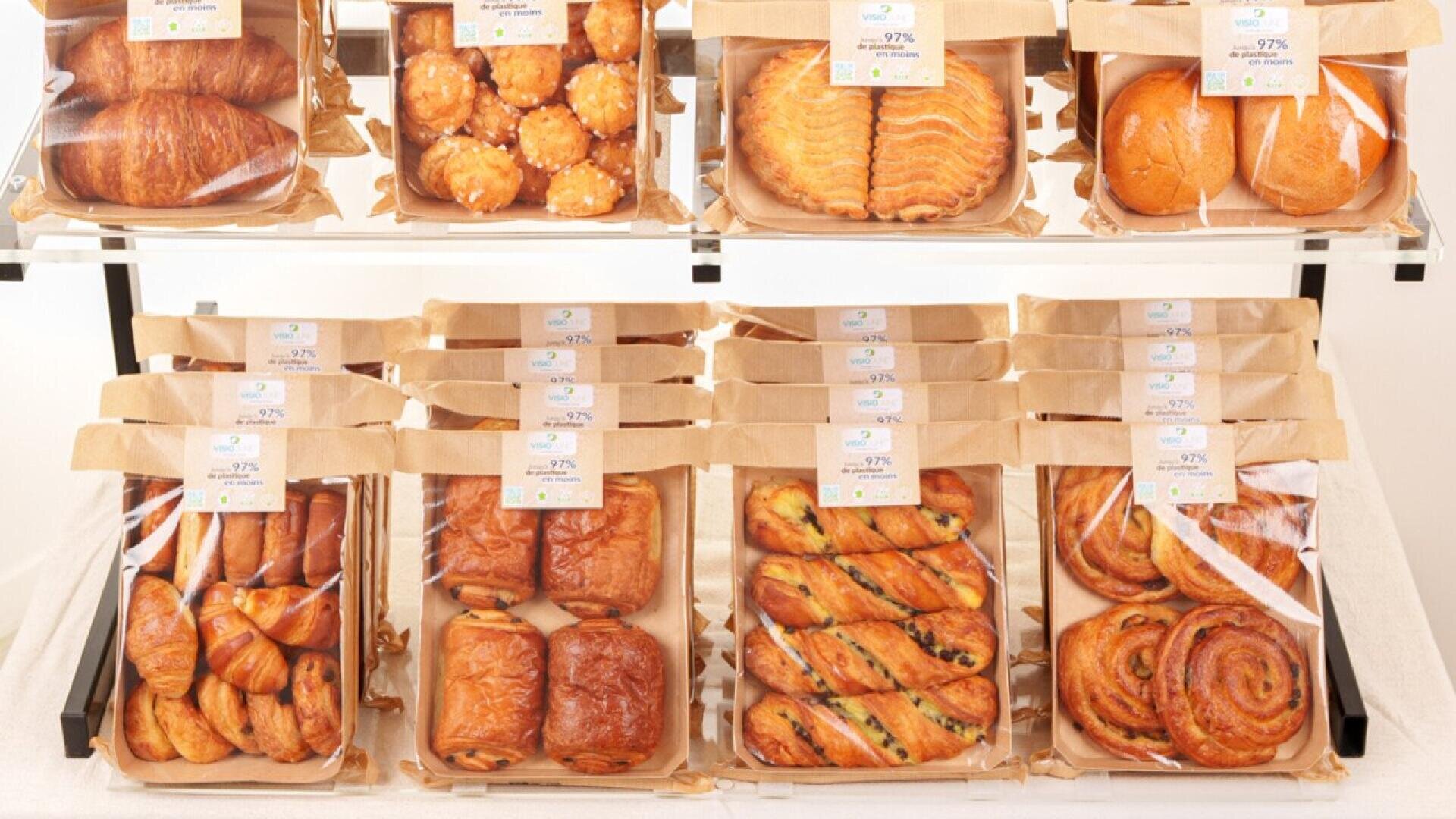 Eine Auslage mit verschiedenen Gebäckstücken, darunter Croissants, Pain au Chocolat, Twists und Swirls, die in Reihen auf Regalen angeordnet sind, jedes in einem braunen Papiertablett mit einer durchsichtigen Abdeckung und einem Etikett obenauf.