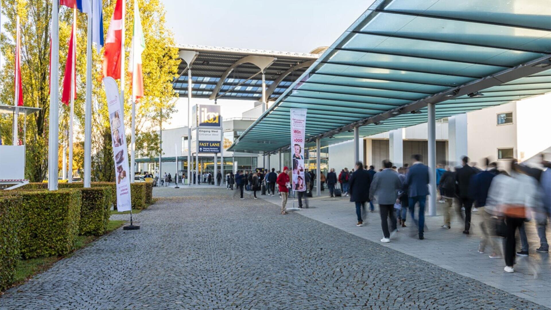 People walk toward a modern building entrance with a glass canopy. Flags and banners line the walkway, and the area is paved with cobblestones. The scene suggests an event or conference.