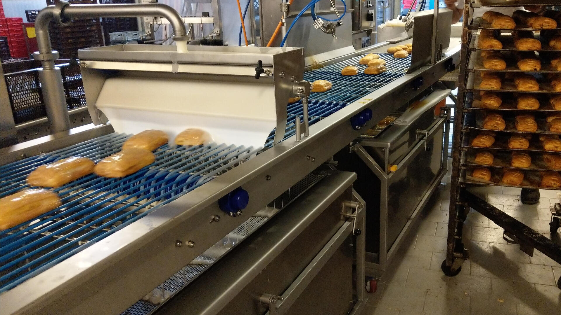 In einer Großbäckerei laufen frisch gebackene Brötchen über ein Fließband, während sich in einer industriellen Umgebung auf einem Regal neben den Maschinen weitere Backwaren stapeln.