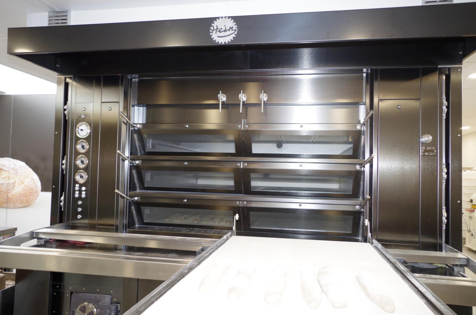 Gezeigt wird ein kommerzieller Brotofen aus Edelstahl mit mehreren Glastüren, vor dem mehrere ungebackene Baguettes auf einem Förderband liegen, bereit zum Backen.