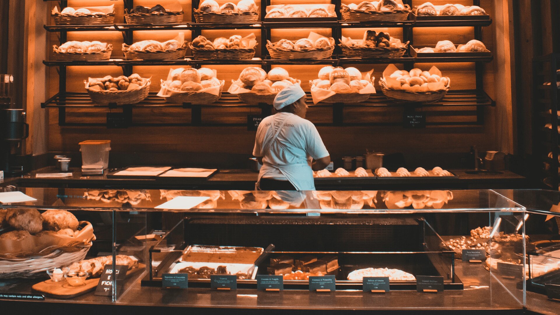 Eine Person in weißer Uniform und mit Hut steht hinter einer Theke in einer Bäckerei, hinter der Regale mit verschiedenen Brotsorten und Backwaren ausgestellt sind. Gebäck und Backwaren sind auch in Tabletts auf der Theke zu sehen.