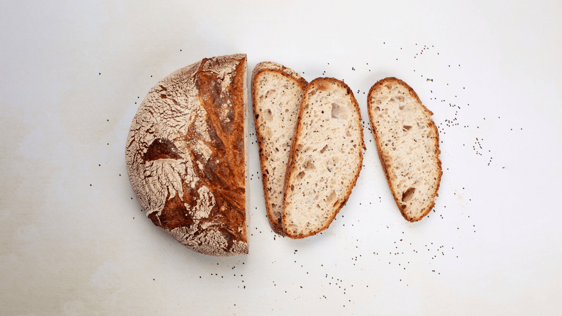 Ein runder Laib rustikalen Brotes, teilweise aufgeschnitten, mit drei Scheiben daneben auf einer hellen Fläche. Einige kleine Krümel sind um das Brot herum verstreut.