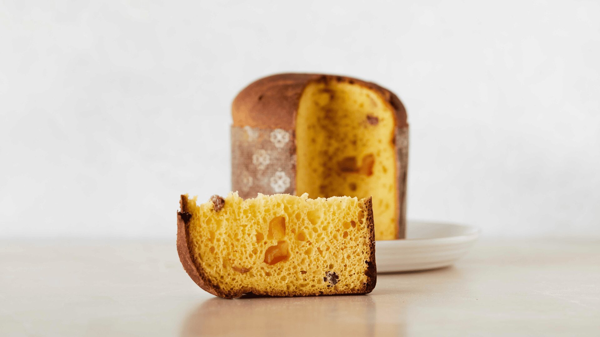 Eine Scheibe Panettone, ein italienisches süßes Brot mit Trockenfruchtstückchen, liegt vor dem restlichen Brot und einem weißen Teller, alles auf einer hellen Fläche mit neutralem Hintergrund.