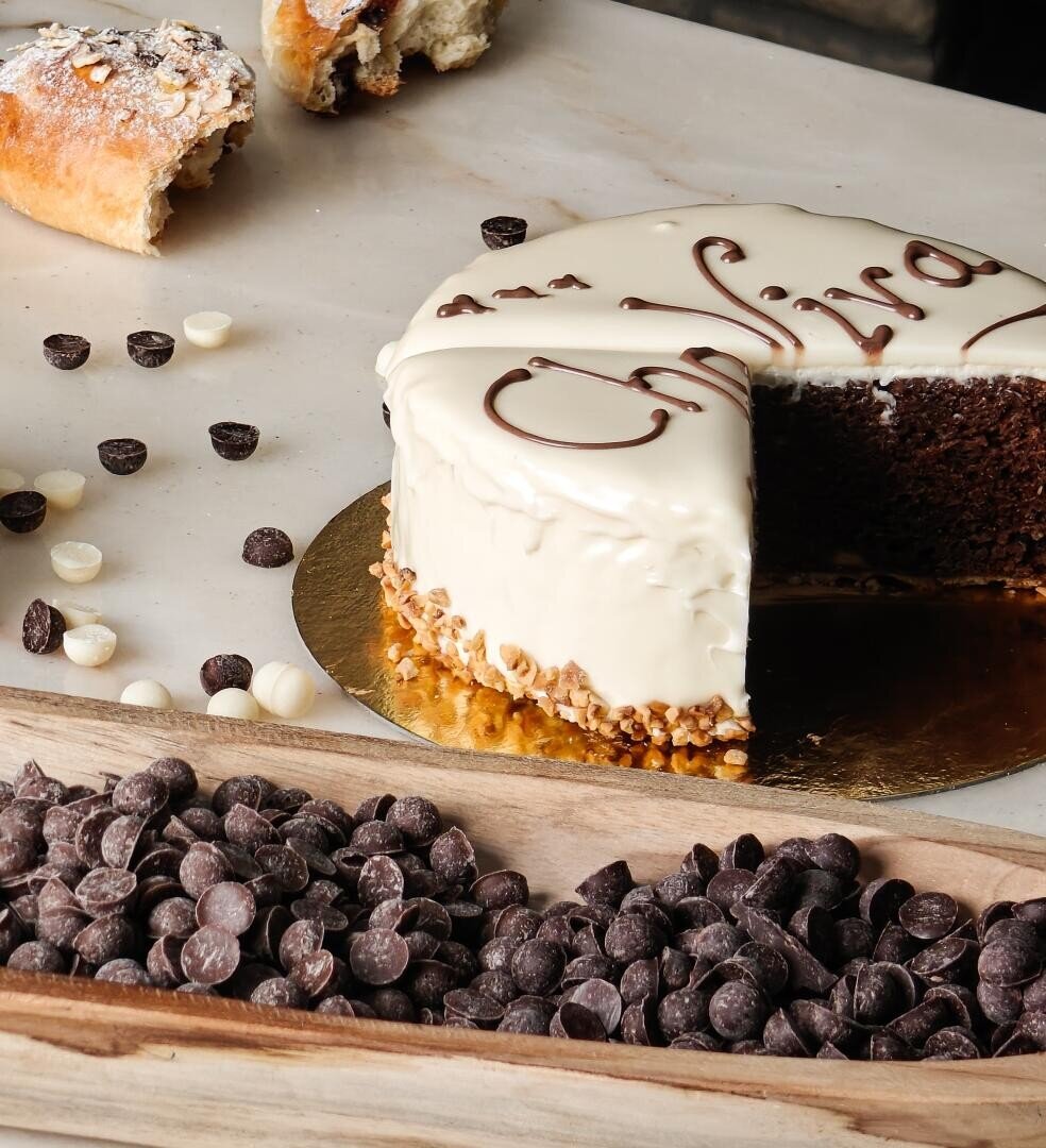 Ein Schokoladenkuchen mit weißem Zuckerguss und dekorativer Schrift, von dem ein großes Stück fehlt, steht auf einer Marmorplatte. Im Vordergrund sind Schokoladenstückchen und ein mit weiteren Stückchen gefülltes Holztablett zu sehen.