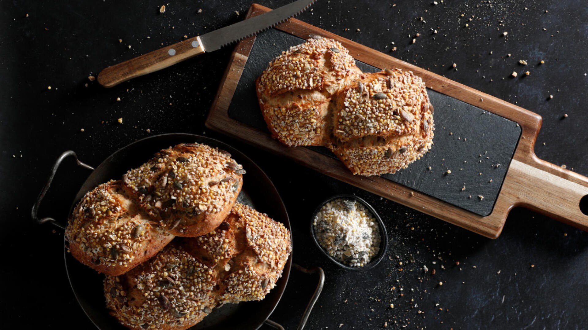 Ein dunkler Tisch mit Körnerbrötchen auf einem schwarzen Tablett, einem Laib Brot auf einem Schneidebrett mit einem Brotmesser und einer kleinen Schale mit Körnern. Krümel und Körner liegen verstreut herum.