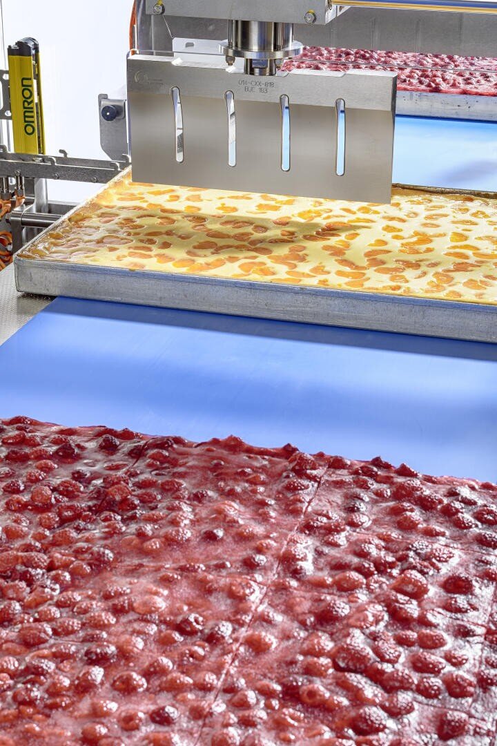 Eine Maschine schneidet Blätterteig mit Erdbeerfüllung auf einem Förderband, wobei die Stücke von einem Tablett auf eine blaue Fläche in einer Lebensmittelverarbeitungsanlage gelangen.