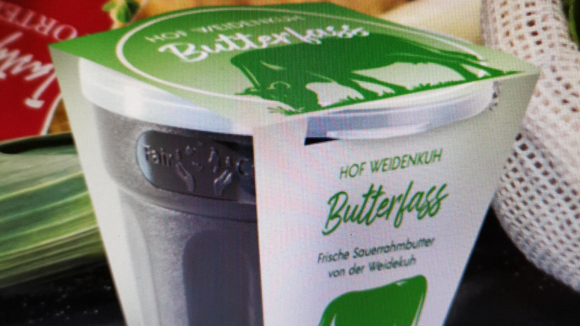 Eine Nahaufnahme eines Behälters Butterfass-Sauerrahm vom Hof Weidenhuh mit grün-weißer Verpackung, einer Kuhillustration und deutschem Text.