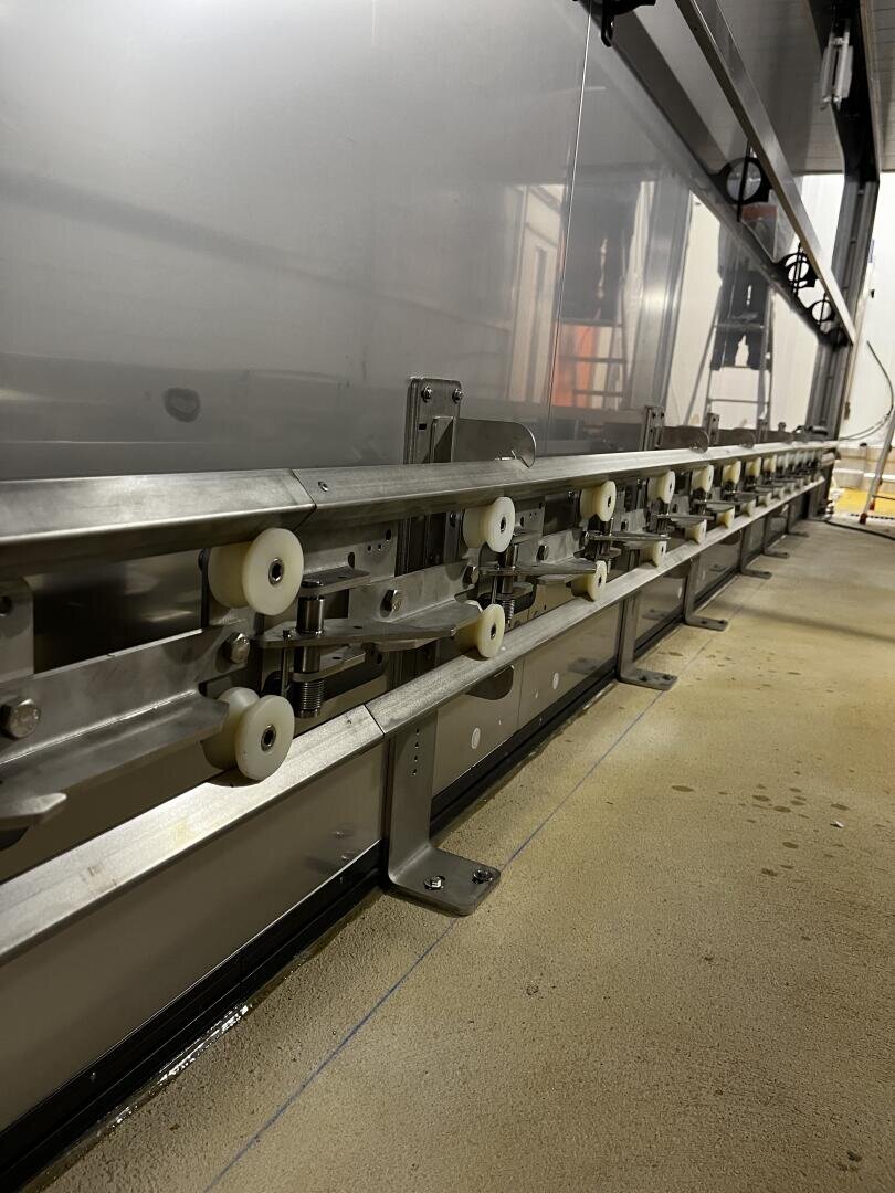 Close-up view of a stainless steel conveyor system with white plastic rollers, mounted on a beige industrial floor next to a metallic wall in a factory or processing facility.