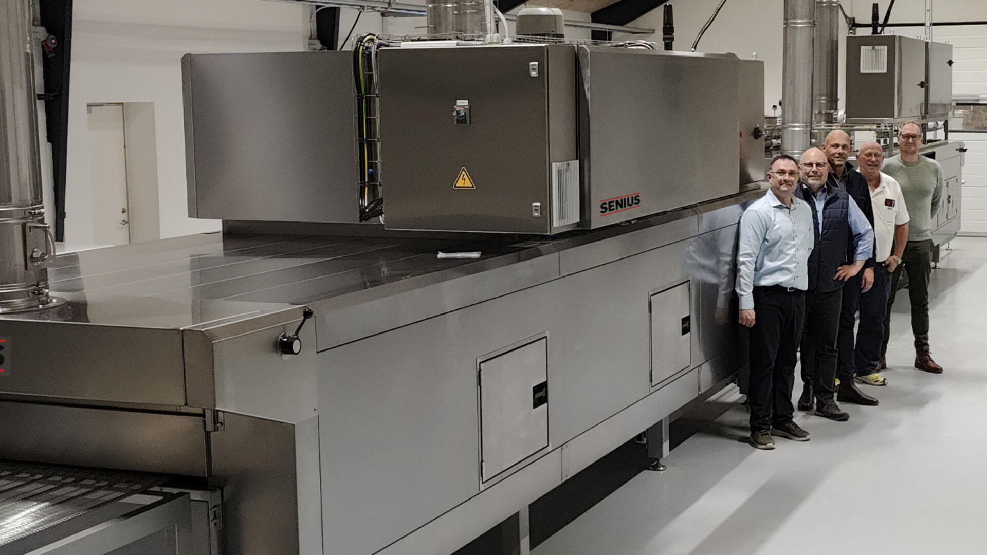 Four men stand beside a large, industrial stainless steel machine inside a modern, clean facility. The machine has the brand name SENIUS on its side and several compartments and control panels.