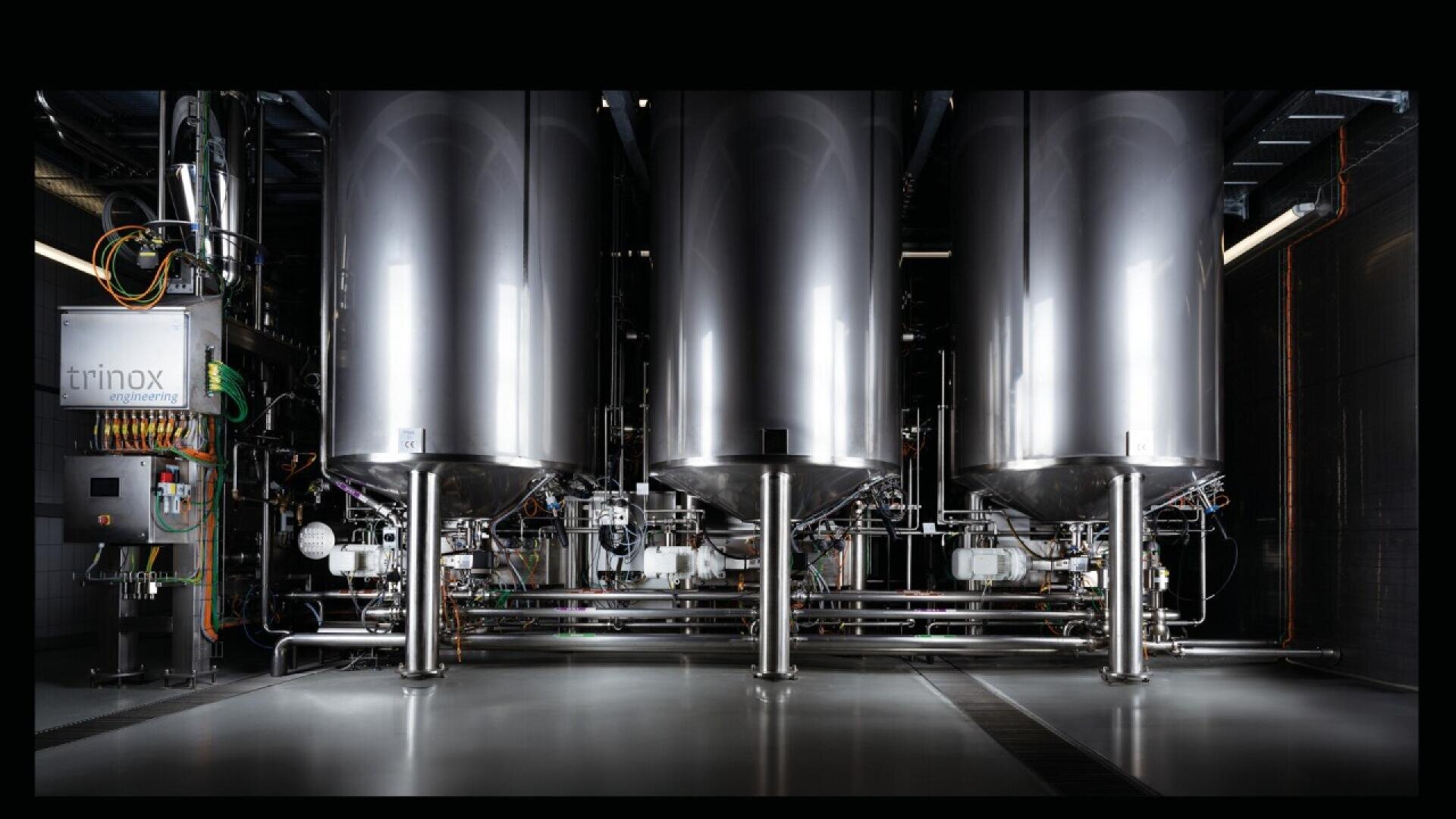 Three large, stainless steel industrial tanks connected by pipes and valves stand in a row in a clean, modern facility. Control panels, including one labeled Trinox, are visible on the left.