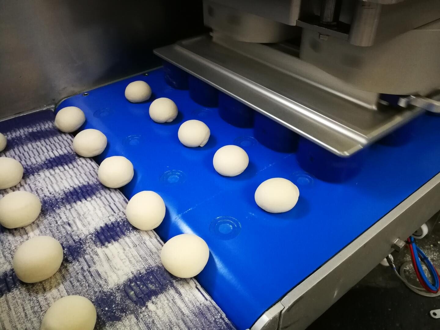 In einer Fabrik werden runde Teigstücke von einem blauen Automaten geformt und angeordnet, während in der Nähe einige Teigkugeln auf einem Förderband liegen.
