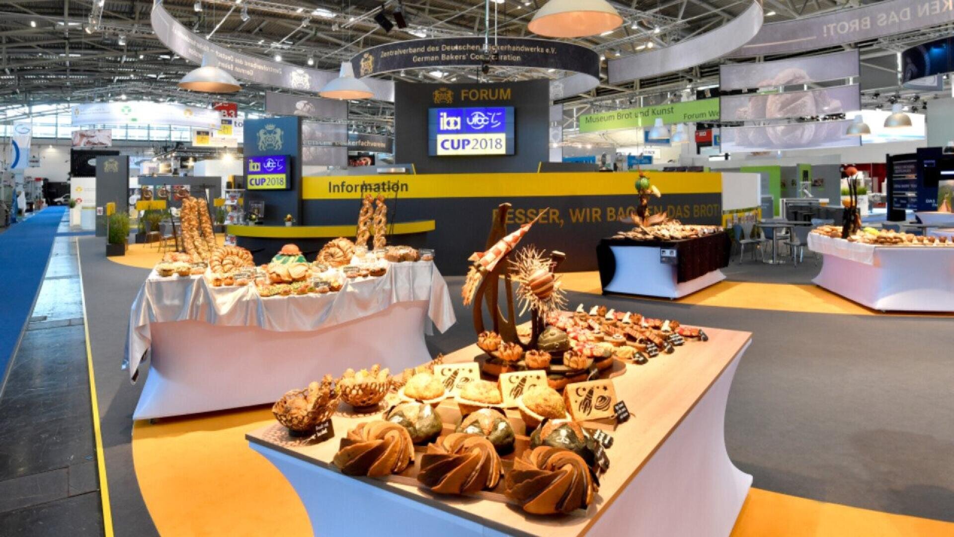 A brightly lit exhibition hall displays various artisan breads and baked goods on decorated tables, with banners, signs, and information booth visible in the background. The setting is lively and spacious.