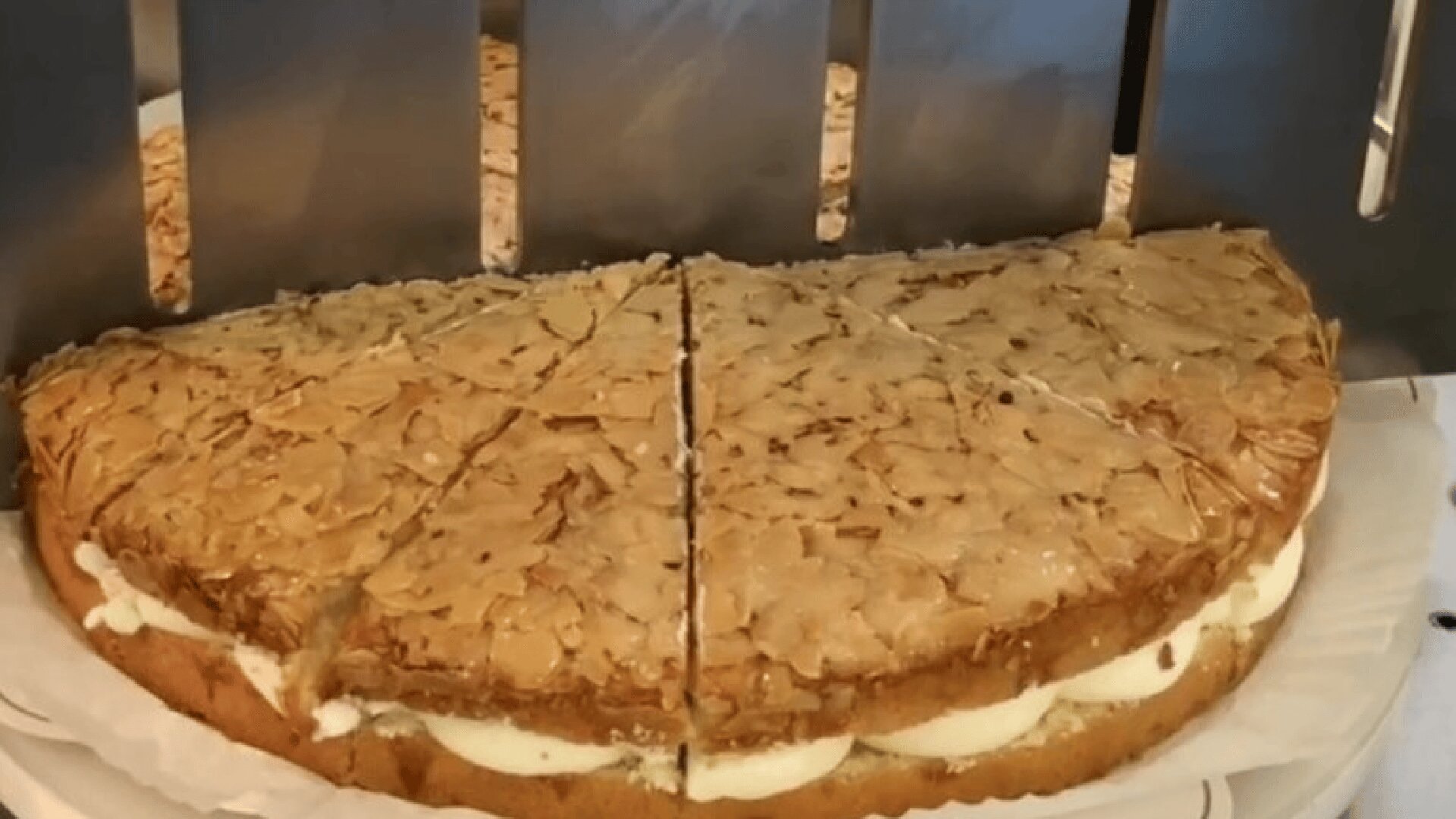 A round cake with a golden, sliced almond topping and a thick layer of cream filling is cut into wedges, sitting on a white tray under a metal slicer.