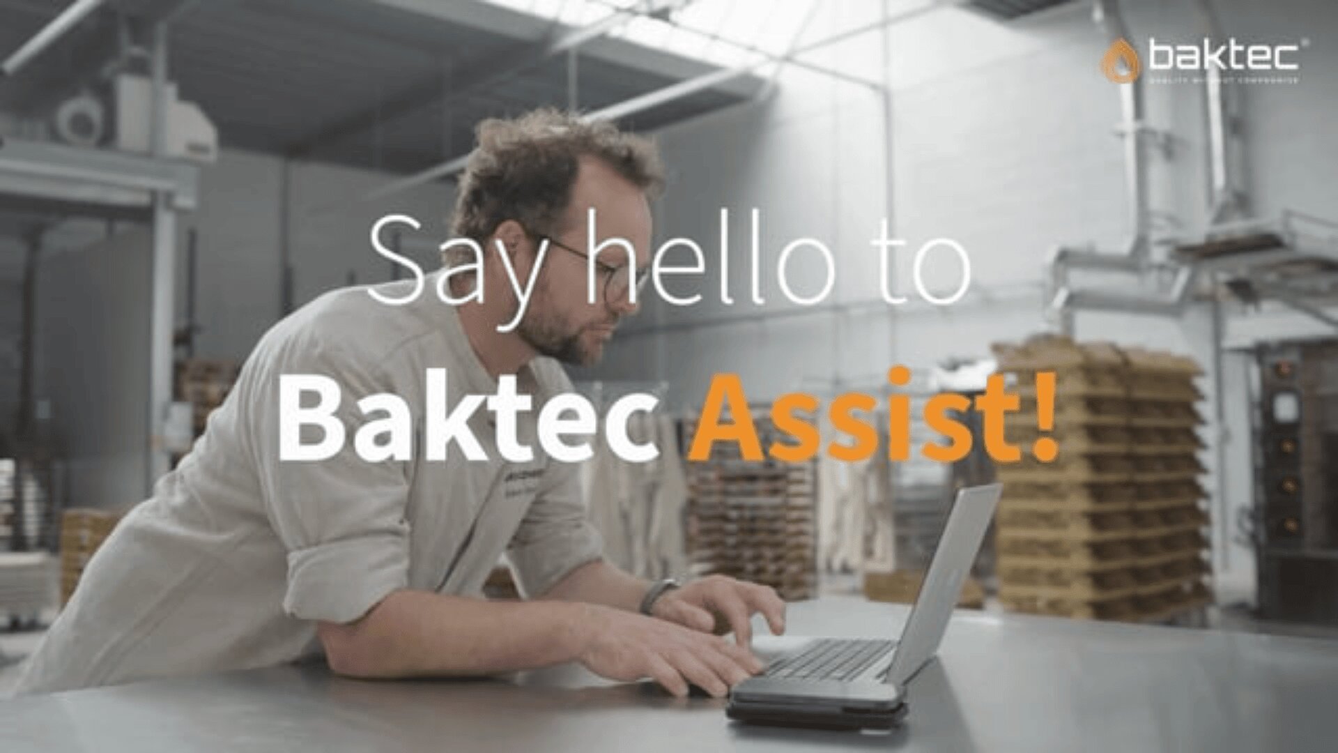 A man in a bakery works on a laptop at a metal table. Stacks of trays are in the background. Overlaid text reads, Say hello to Baktec Assist! with the Baktec logo in the top right corner.