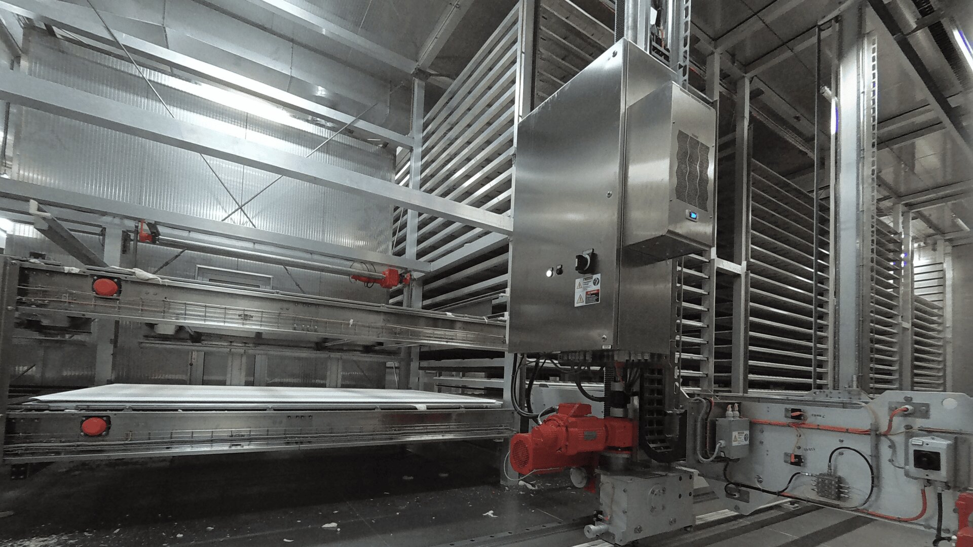 Large industrial food processing equipment with multiple stacked metal trays and conveyor belts inside a bright, clean factory setting. Control panels and red machine parts are visible in the foreground.