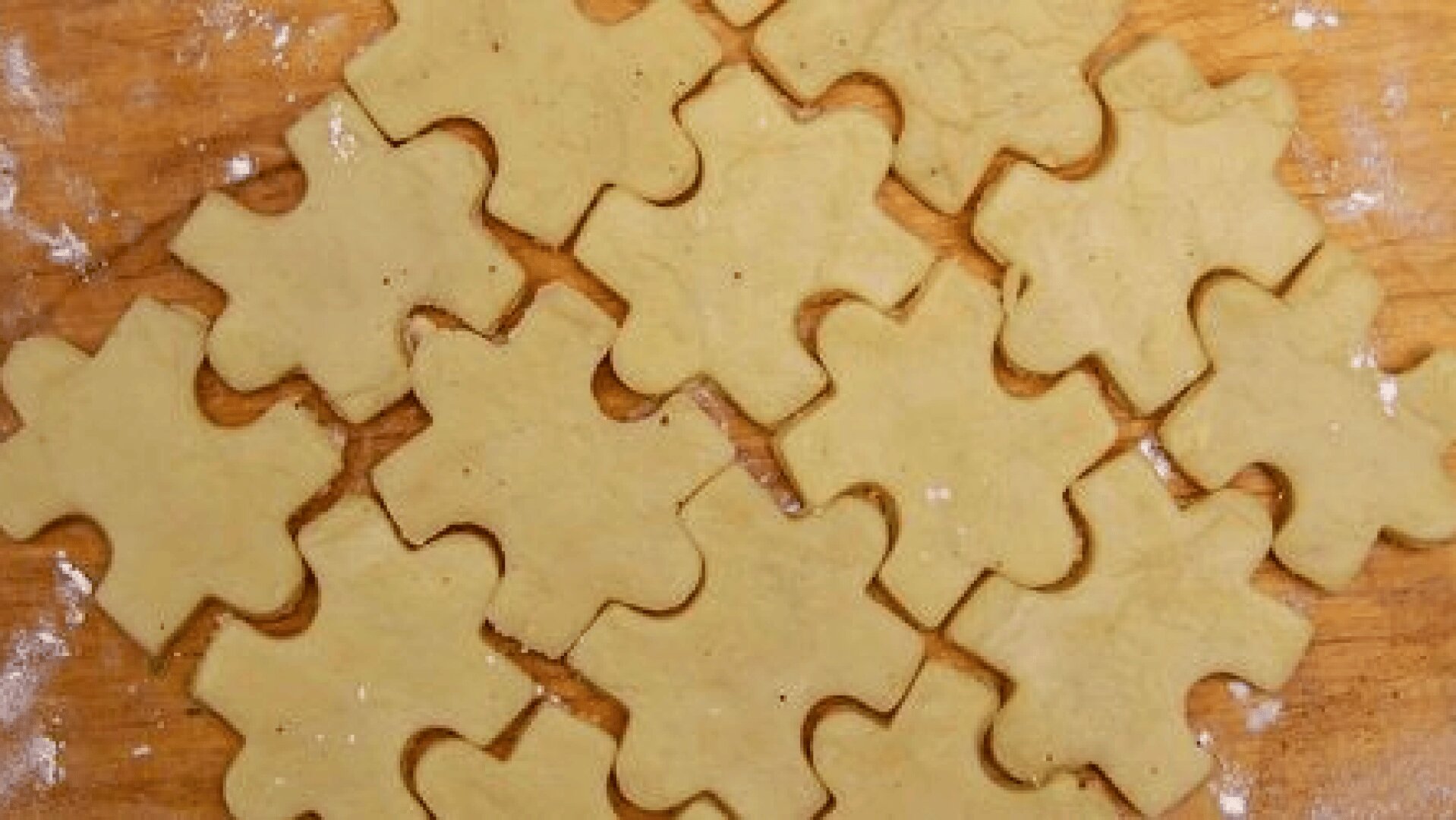 Puzzle-shaped cookie dough pieces are arranged together on a floured wooden surface, ready to be baked.