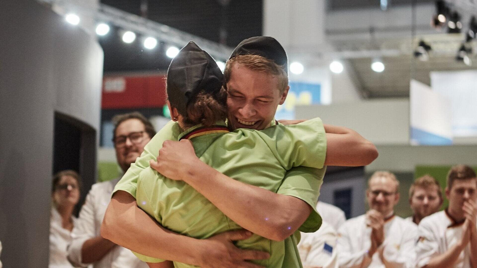 Two people in chef uniforms hug joyfully in a brightly lit room, while others in similar attire stand in the background clapping and smiling, suggesting a celebratory or competition setting.